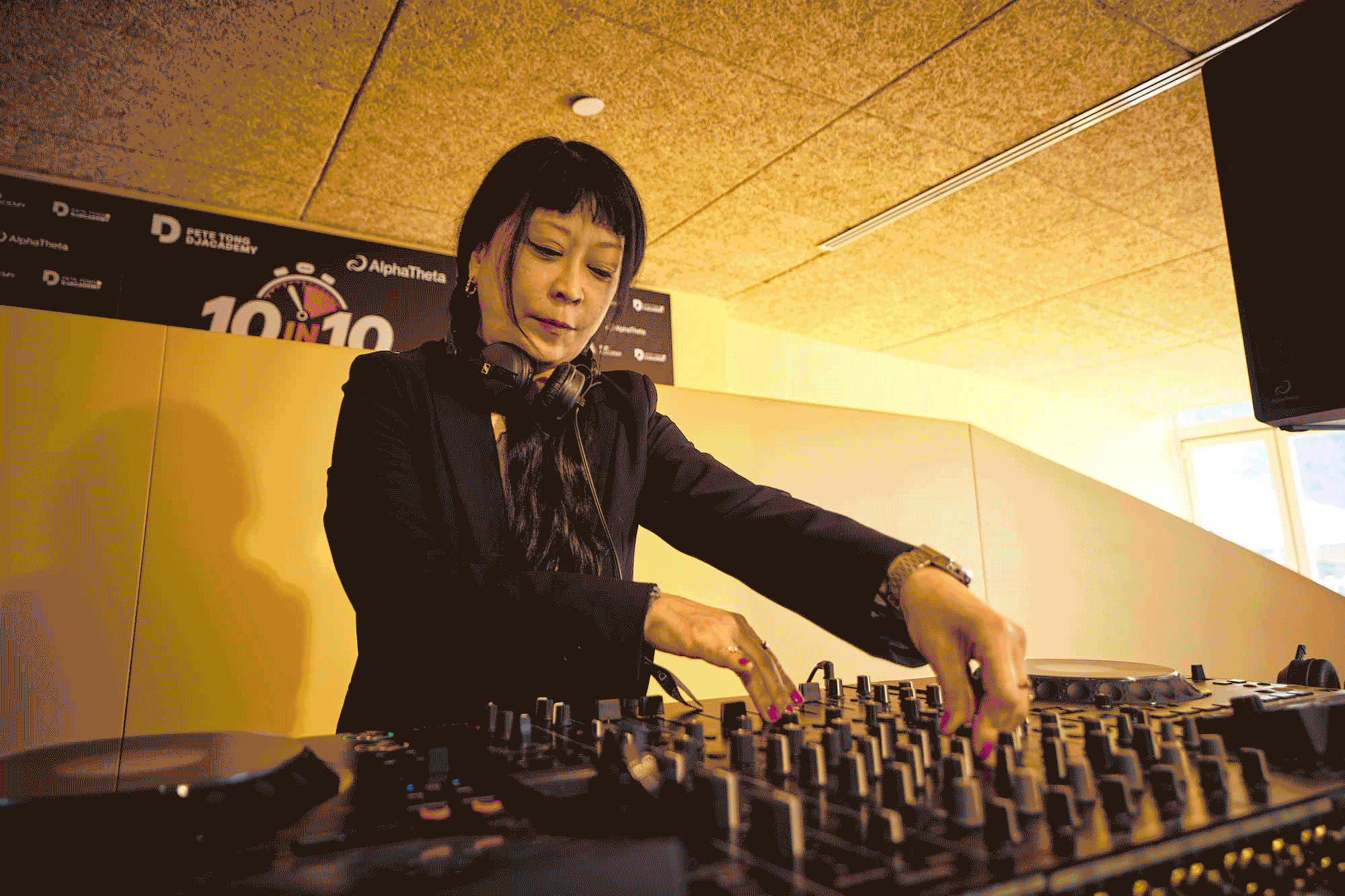 Female DJ wearing headphones around her neck and adjusting mixer knobs in a warmly lit studio, concentrating on the music.