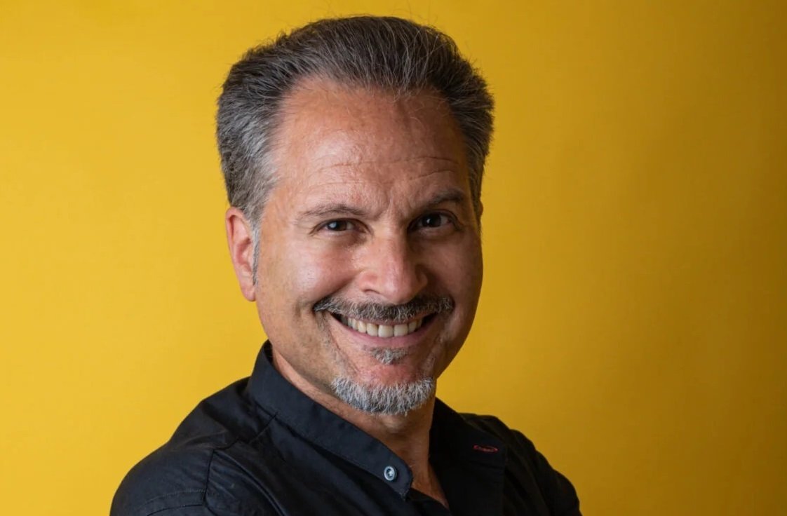 Smiling middle-aged man with gray hair and a goatee against a solid yellow background.