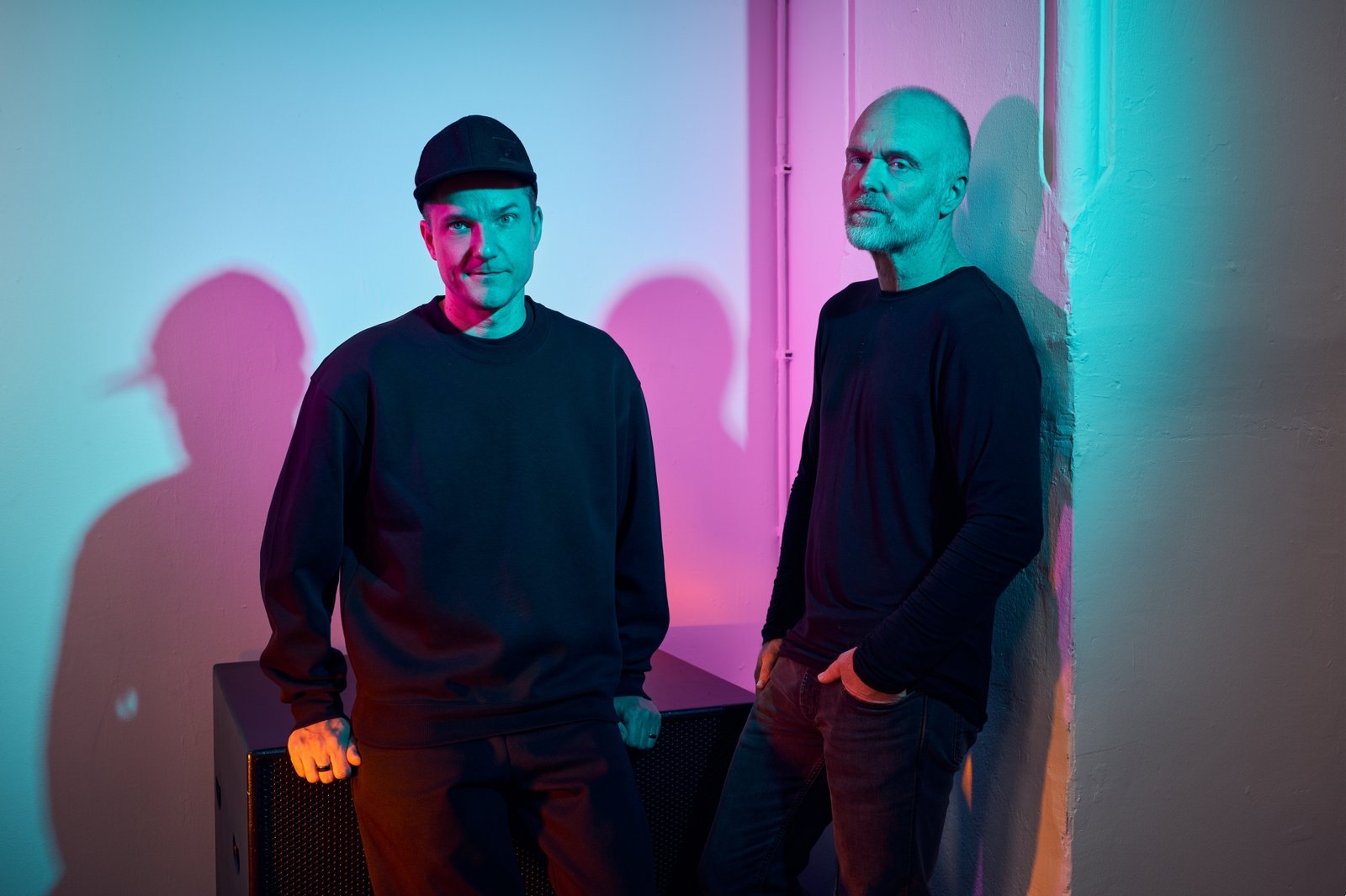Two men in dark clothing pose in a studio with teal and pink lighting; one wears a cap, leaning near an amplifier.
