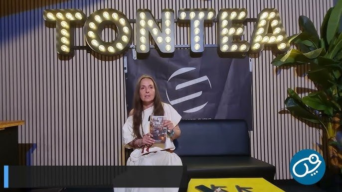 Woman seated on a black couch in a studio, holding a magazine with large illuminated 'TONTEA' sign behind her.
